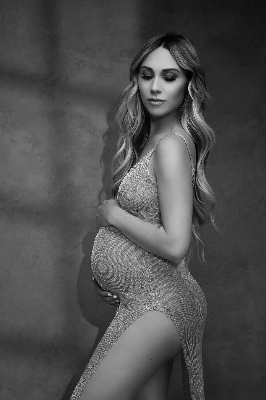 maternity portrait of a pregnant woman standing by a wall with a window light pattern behind her while wearing a sheer dress taken by Andrea Liora Studios in San Francisco CA