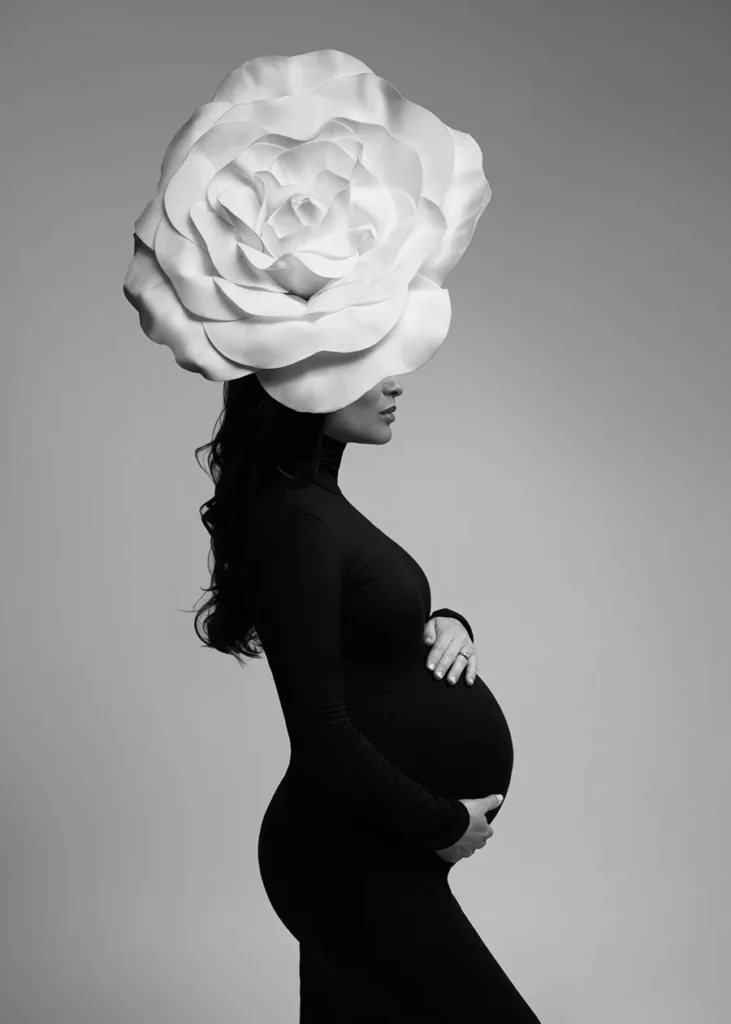 Black and white maternity photo of a woman in a black turtleneck dress and white flower crown on her head. The flower covers her face making the maternity photograph artistic in feeling.