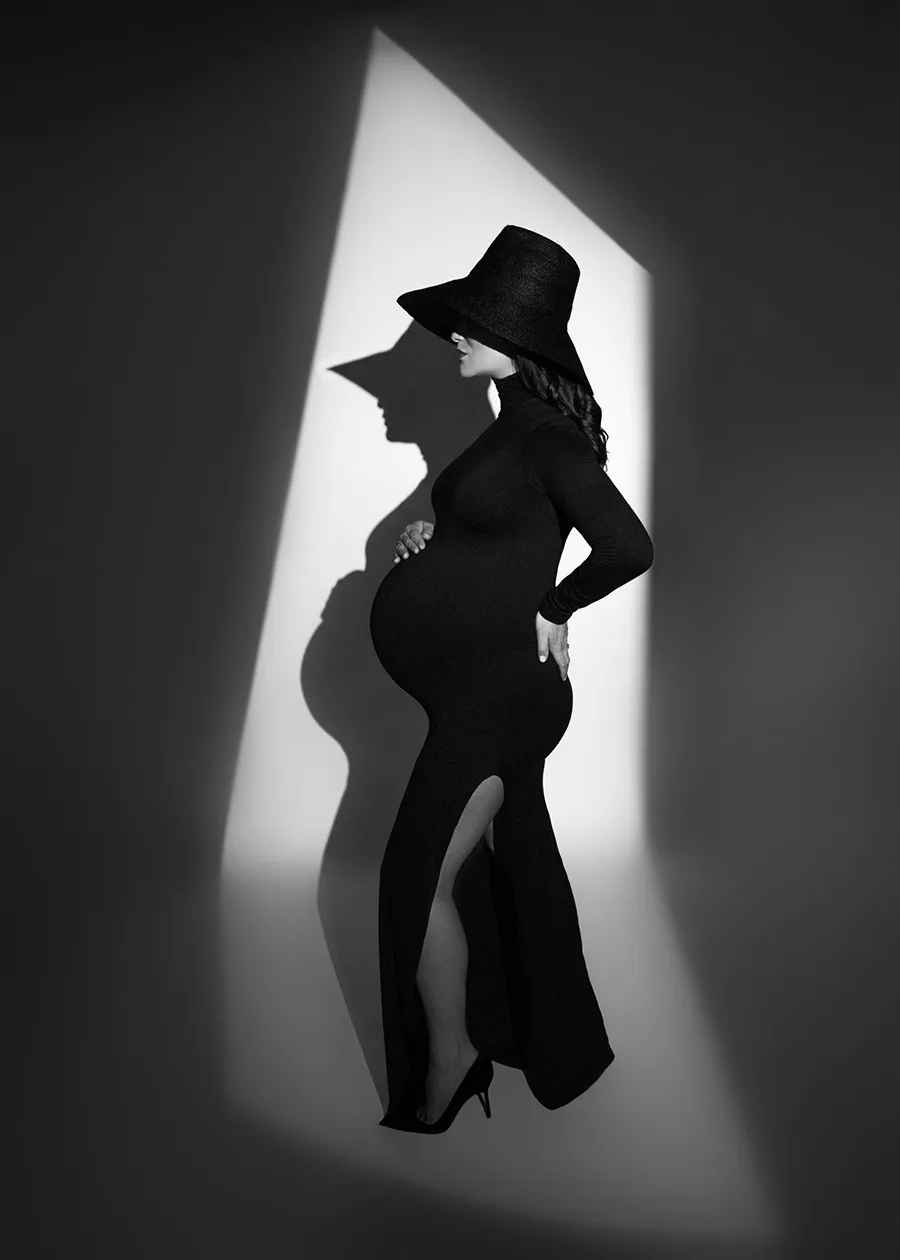Black and white maternity photo of a woman in a black turtleneck dress and black wide brimmed hat on her head. The hat covers her face making the maternity photograph artistic in feeling.