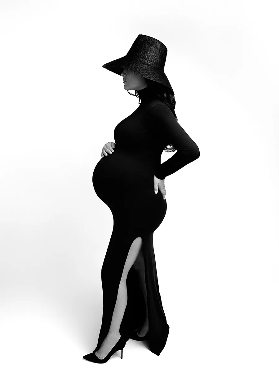 Black and white fashion style maternity photo of a woman in a black turtleneck dress and black wide brimmed hat on her head. The hat covers her face making the maternity photograph artistic in feeling.
