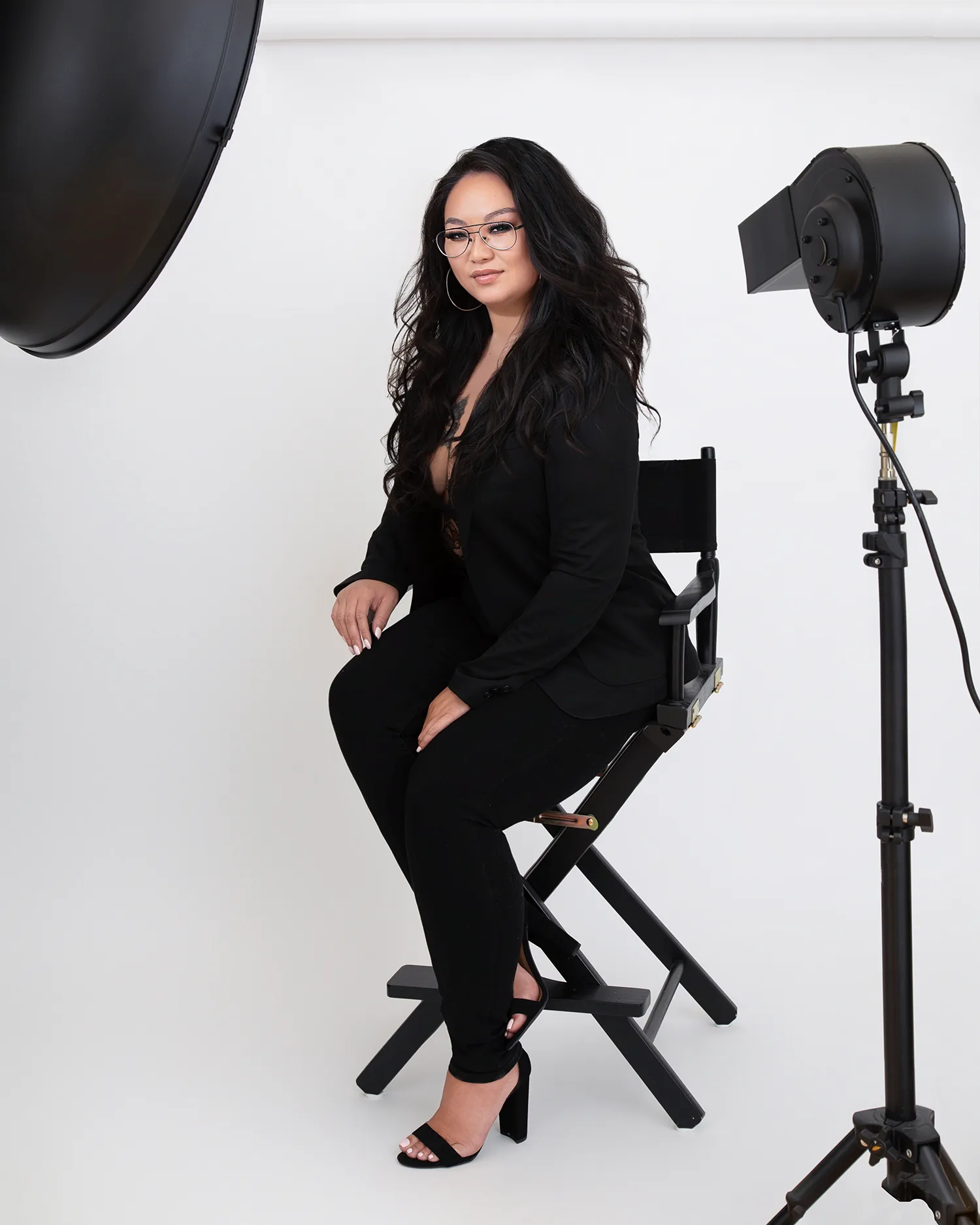 Portrait of a woman in a black suit sitting in a black directors chair with a fan machine blowing wind in her hair and the corner of a beauty dish light in the foreground.