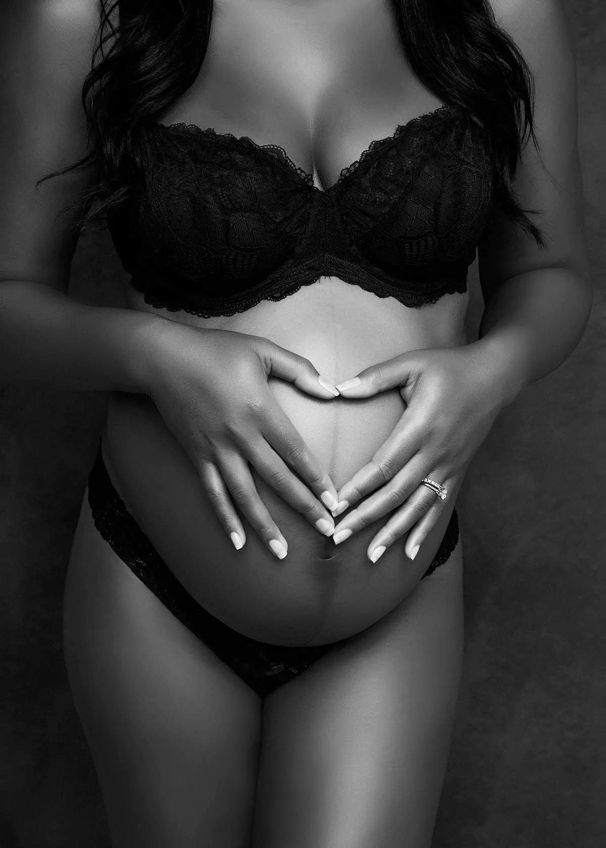 Black and white artistic boudoir maternity portrait of a pregnant woman wearing a black bra and pant. Image is focused on her belly with her hands making a heart shape on top. Maternity photograph by Andrea Liora Studios in San Francisco CA.